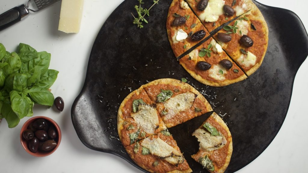 2 small pizzas on pizza plate, Cheese, Olives and Basil on table.